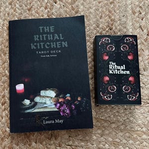 May include: A book and a tarot deck with a dark cover. The book is titled "The Ritual Kitchen" with the subtitle "Tarot Deck Food, folk, fortune" and the author's name "Laura May". The tarot deck has a matching design.