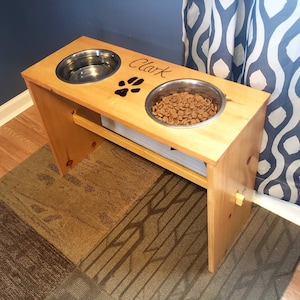 May include: A wooden dog food and water stand with two stainless steel bowls. The stand is stained a light brown color and has the name "Clark" engraved on the top. A paw print is also engraved on the top of the stand.