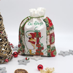Puede incluir: Una bolsa de regalo de Navidad blanca con un diseño de patchwork verde y rojo. La bolsa presenta un pato con un gorro de Papá Noel y sosteniendo un regalo. El texto "De Santa" está bordado en la bolsa.