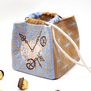 May include: A blue and brown fabric tote bag with a white drawstring. The bag has an embroidered design of a clock with gears and a keyhole.