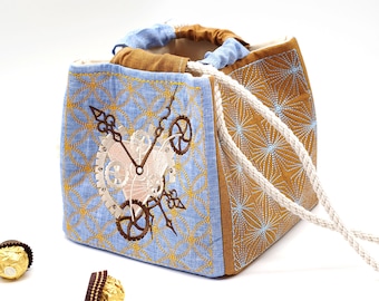 May include: A blue and brown fabric tote bag with a white drawstring. The bag has an embroidered design of a clock with gears and a keyhole.