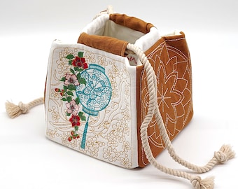 May include: A square, open-top drawstring bag with a floral design. The bag features a white background with embroidered flowers and a teal lantern design. The sides are brown with a stitched floral pattern, and the drawstring is a thick, off-white rope.