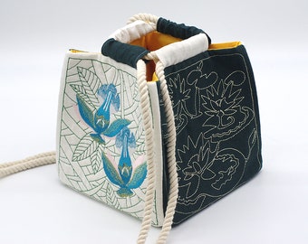 May include: A white and dark green fabric tote bag with a rope handle. The bag has embroidered floral designs on both sides. One side features blue and pink flowers, while the other side features a green and white floral design.
