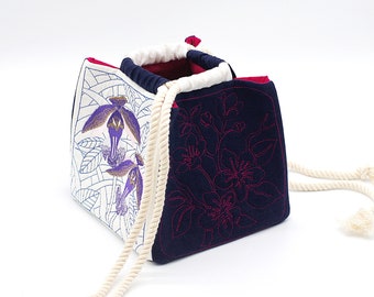 May include: A white and navy blue drawstring bag with floral embroidery. The bag has a white rope drawstring and is made of a soft fabric.