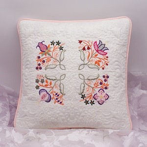 Puede incluir: Una almohada blanca con un borde rosa presenta un diseño floral bordado en tonos naranja, rosa, morado y verde. El diseño está centrado en la almohada y presenta un patrón repetitivo de flores y hojas.
