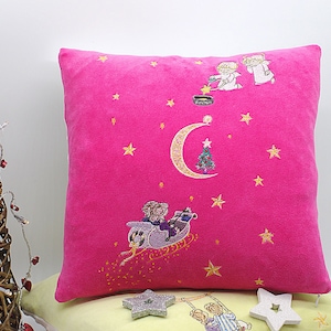 May include: Pink velvet pillow with embroidered Christmas scene featuring angels, a crescent moon, a Christmas tree, and a sleigh with two figures. The background is a dark pink with gold stars.