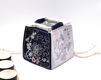 May include: A square, fabric tote bag with a white interior and a navy blue exterior. The navy blue side features white and pink floral embroidery. The other side is light pink with blue bird and floral embroidery. The handle is made of a thick, white rope.