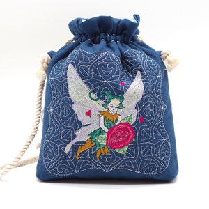 Puede incluir: Una bolsa azul con cordón y un diseño bordado detallado. El diseño presenta un hada sosteniendo una rosa roja, con detalles bordados en blanco y un borde decorativo. La bolsa tiene un cordón de cuerda color crema.