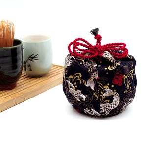 May include: A black fabric drawstring pouch featuring a gold and white koi fish pattern, secured with a red cord. Beside it sits a bamboo whisk in a dark green ceramic cup and a light green ceramic cup on a rectangular bamboo tray.
