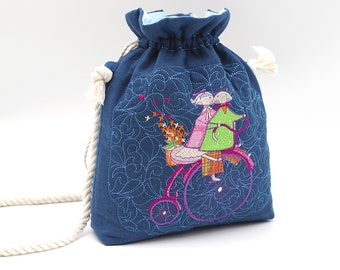 Puede incluir: Una bolsa de cordón azul con un asa de cuerda blanca. La bolsa presenta un diseño bordado de dos figuras montando una bicicleta con flores.