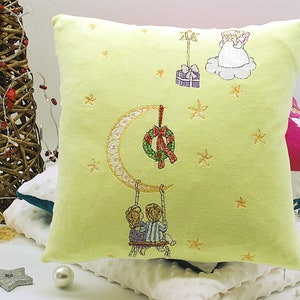 May include: A yellow pillow with embroidered Christmas designs. The pillow features a crescent moon with a wreath, two children on a swing, a star, and an angel with a gift.