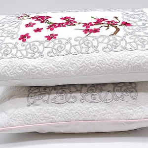 Puede incluir: Dos almohadas decorativas blancas con ribete rosa. La almohada superior presenta un diseño floral bordado gris con flores rosas. La almohada inferior tiene un diseño bordado gris similar.