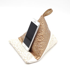 May include: A white and gold phone stand with a quilted design. The stand has a handle and is designed to hold a smartphone upright.
