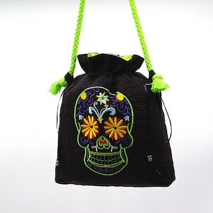 以下が含まれることがあります： 緑色のロープハンドルとカラフルな刺繍のシュガースカルデザインが施された黒い巾着袋です。