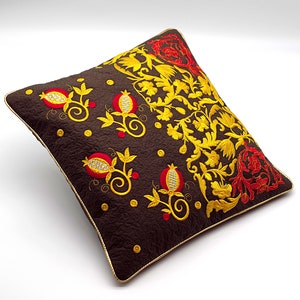 Puede incluir: Una almohada marrón con diseños florales bordados en oro y rojo. La almohada presenta un patrón repetitivo de granadas y hojas.