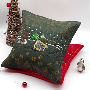 Puede incluir: Dos cojines navideños decorativos. El cojín superior es verde oscuro con estrellas bordadas, copos de nieve y una escena invernal. El cojín inferior es rojo con un diseño de estrella. Un pequeño adorno de pingüino y un árbol decorativo están en el fondo.