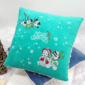 May include: A teal blue throw pillow with white embroidered snowflakes and the text "Merry Christmas". The pillow features two embroidered images of cartoon dogs pulling a sled and a snowman with a child.