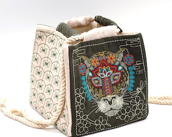 May include: A green and white canvas tote bag with an embroidered design of a colorful animal face. The animal has a floral headdress and a white background.