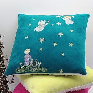 May include: A teal blue throw pillow with embroidered angels and stars. The pillow is layered on top of a yellow pillow with a white border.