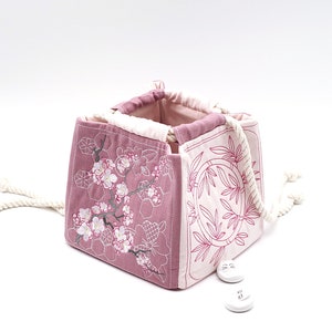 Puede incluir: Una bolsa de tela rosa con un diseño floral y un asa de cuerda blanca. La bolsa está hecha de tela de patchwork con diferentes diseños bordados.