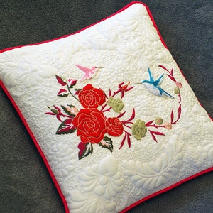 Puede incluir: Almohada blanca con borde rojo, con rosas rojas bordadas, hojas verdes y dos colibríes, uno rosa y otro azul.