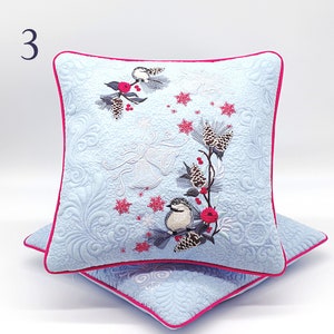 Peut inclure: Deux coussins carrés décoratifs bleu clair avec un motif matelassé et une bordure rose. Chaque coussin présente un motif brodé d'oiseaux, de pommes de pin et de baies rouges. Le chiffre "3" est dans le coin supérieur gauche.