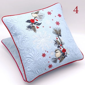 May include: A light blue square throw pillow with red piping, featuring embroidered chickadees on pine branches with red berries, pinecones, and white snowflakes. The fabric has a quilted texture with swirling patterns and the number 4.