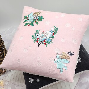 May include: A pink square pillow with embroidered angels and snowflakes. The angels are in light blue and white, with holly and red berries. The pillow has a white scalloped edge and is stacked on other pillows.