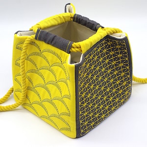 May include: A yellow fabric drawstring bag with a geometric pattern on one side and a wave pattern on the other side. The bag has a thick yellow rope handle.