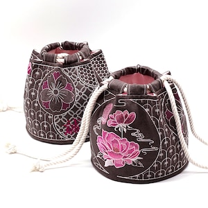 Könnte beinhalten: Zwei braune Stoffbeutel mit rosa Blumenstickerei. Die Beutel haben weiße Kordeln und sind mit einem geometrischen Muster verziert.