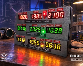 May include: A silver-toned electronic time display unit featuring three stacked digital LED panels. The top panel displays red text for Destination Time, the middle panel shows green text for Present Time, and the bottom panel displays yellow text for Last Time Departed, each with month, day, year, hour, and minute indicators.