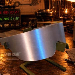 Op de afbeelding: Een gebogen, zilverkleurig metalen vizier met een glad, reflecterend oppervlak, ondersteund door een groene plastic standaard. Het vizier heeft een opvallende uitsparing langs de onderrand en wordt getoond in een donkere werkplaatsomgeving.
