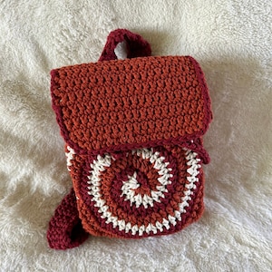 May include: A small, crocheted backpack with a red and white circular pattern on the front. The backpack has a red strap and is made with a textured yarn.
