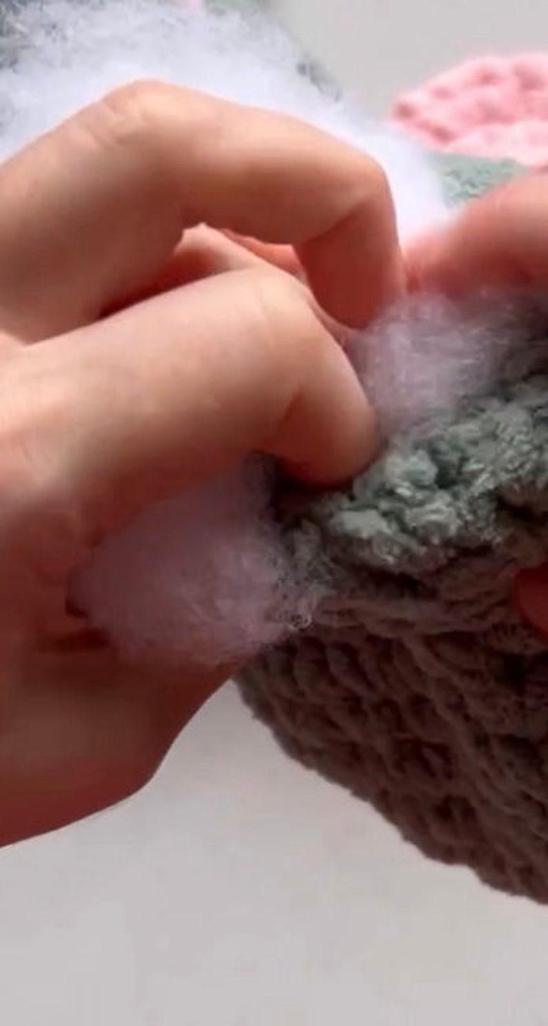 May include: A close-up of a hand holding a multi-colored knitted hat. The hat is made of soft, fluffy yarn in shades of pink, green, and gray.