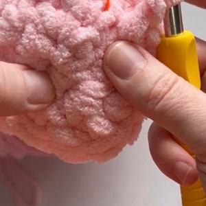 May include: Close-up of a person crocheting with a pink yarn and a yellow crochet hook. The yarn is fluffy and the hook is metal with a yellow handle.