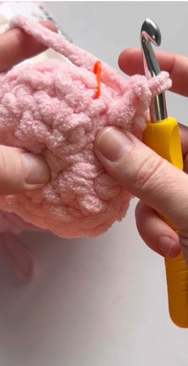 May include: Close-up of a person crocheting with a pink yarn and a yellow crochet hook. The yarn is fluffy and the hook is metal with a yellow handle.