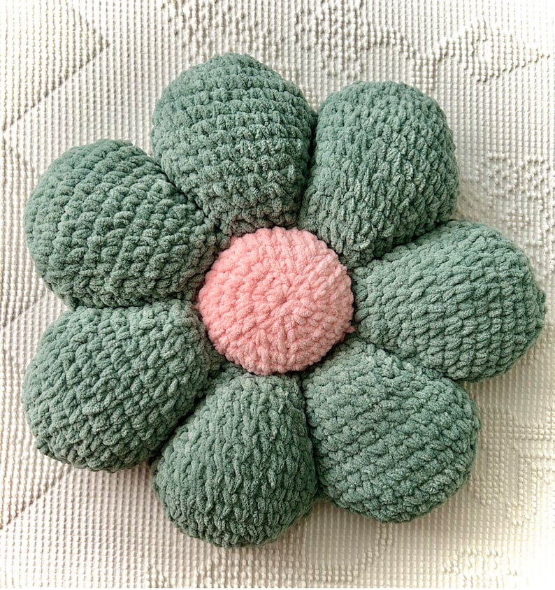 May include: A green and pink flower-shaped pillow with a textured fabric. The pillow is made of multiple fabric pieces sewn together to form a flower shape.