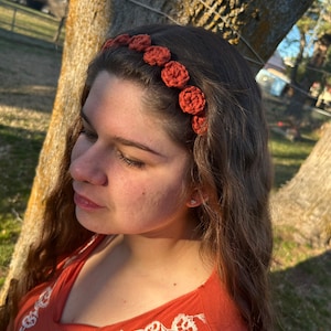 May include: A woman with long brown hair wearing a terracotta crocheted headband with a floral design. She is wearing a rust-colored top with white embroidery.
