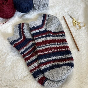 May include: A pair of striped knitted socks in grey, navy blue, and burgundy. The socks are lying on a white fluffy surface.