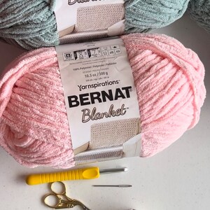 May include: Two skeins of yarn, one pink and one light blue, labeled "Bernat Blanket" yarn. The yarn is 100% polyester and weighs 10.5 ounces or 300 grams. The yarn is for crocheting or knitting blankets. There are also knitting needles, scissors, and a small yellow fabric container with colorful yarn scraps.