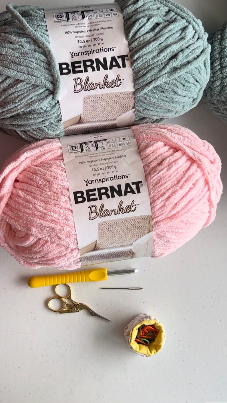 May include: Two skeins of yarn, one pink and one light blue, labeled "Bernat Blanket" yarn. The yarn is 100% polyester and weighs 10.5 ounces or 300 grams. The yarn is for crocheting or knitting blankets. There are also knitting needles, scissors, and a small yellow fabric container with colorful yarn scraps.