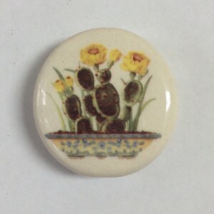 May include: A round, off-white ceramic tile featuring a botanical illustration. The design showcases a cactus with yellow flowers and green leaves, planted in a decorative blue and yellow pot. The tile has a glossy finish.