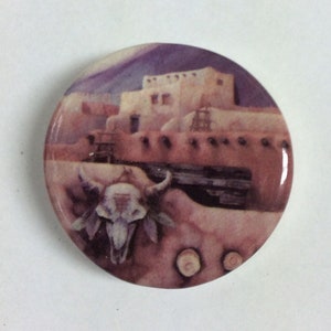May include: A round ceramic coaster with a painted image of a Southwestern adobe building with a buffalo skull in the foreground. The background is a purple and pink sunset.