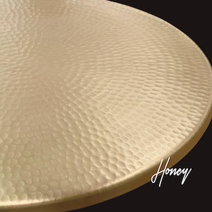May include: A circular, light gold-colored tray or plate featuring a distinct hammered texture across its entire surface. The metal surface has a matte finish with a repeating dimpled pattern. The word Honey is printed in white script in the corner.