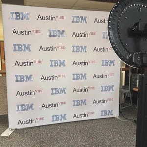May include: A photo booth setup with a white backdrop displaying the logos "IBM" and "Austin VIBE" in blue. The booth includes a large, circular light and a black stand. The backdrop is set against a grey carpet.