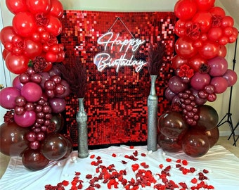 Könnte beinhalten: Geburtstagsdekoration mit roten und weinroten Luftballons, Blumenschmuck und einer Paillettenwand. Die Worte "Happy Birthday" sind in weißem Neon gehalten. Dekorative Vasen und verstreute Rosenblätter vervollständigen die Szene.