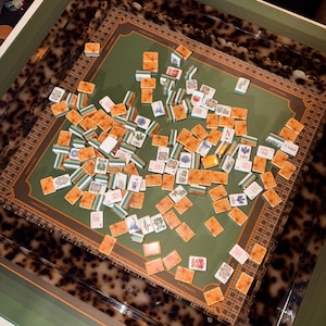 Op de afbeelding: Een Mahjong-set met een groen speeloppervlak en een rand met schildpadpatroon. De set bevat talrijke tegels met verschillende symbolen en tekens, verspreid over het bord. Een doos met tekst is linksboven zichtbaar.