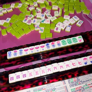 May include: A Mahjong set with green and white tiles, arranged on a pink surface. The tiles display various symbols and numbers. The set includes racks with numbers and symbols, and a chart with text.