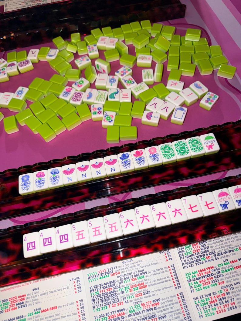 May include: A Mahjong set with green and white tiles, arranged on a pink surface. The tiles display various symbols and numbers. The set includes racks with numbers and symbols, and a chart with text.