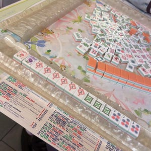 May include: A Mahjong set with white and orange tiles, arranged on a floral patterned surface. The tiles display various symbols and numbers, with a clear plastic tray for the tiles and a printed game guide.